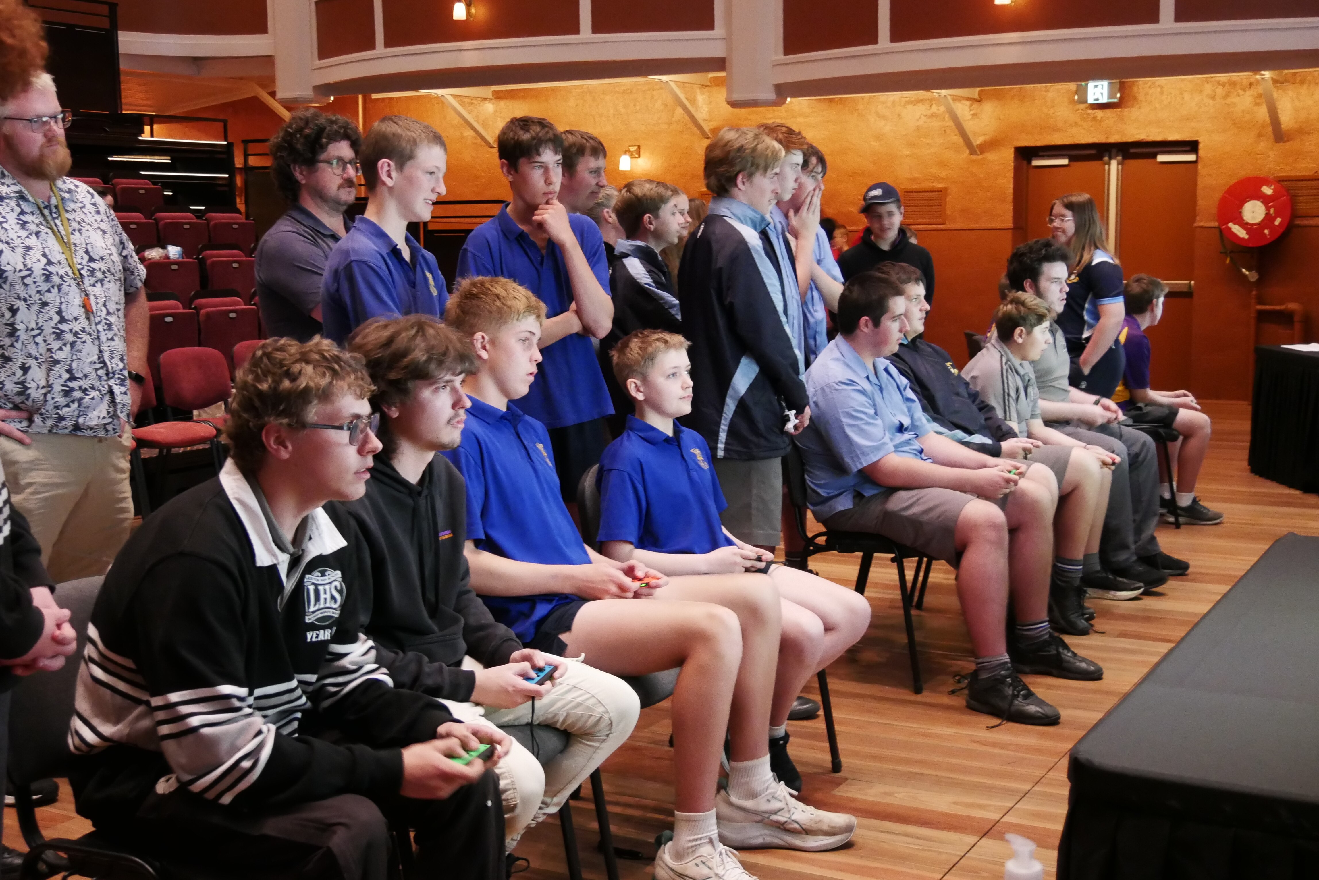 Eight boys sit on chairs in front of two tv screens playing a game while a crowd stands behind them. 