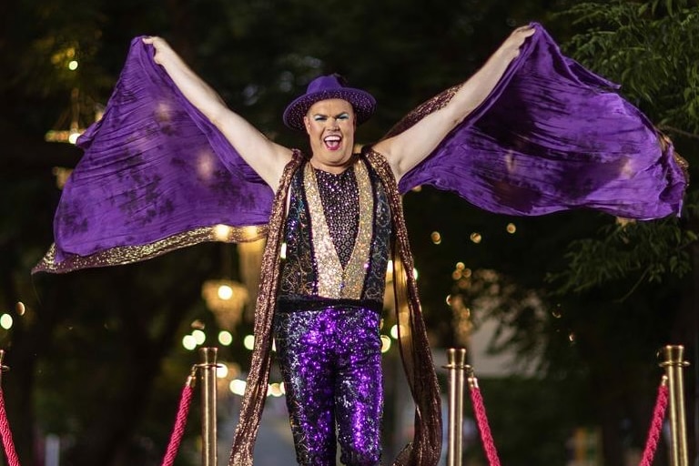 man in purple sequin outfit with bare arms raised in victory smiling