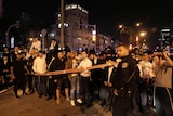 A crowd of men stands behind police tape at night