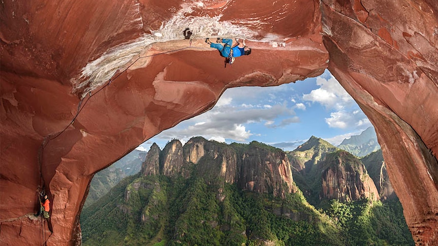 A rock climber crawls upside down along an overhang, with a beautiful vista of mountains in the background.