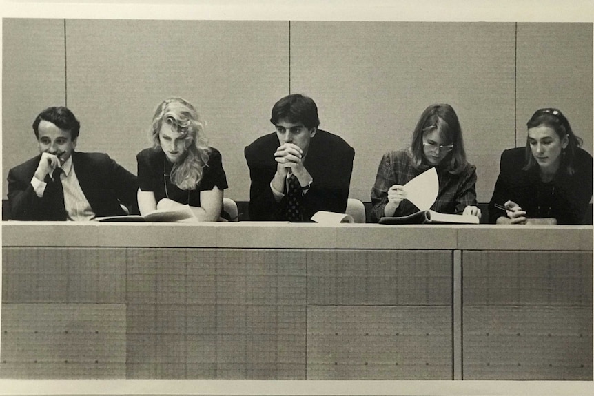 Black and white photo of five journalists in press gallery looking down at documents or at floor of parliament.