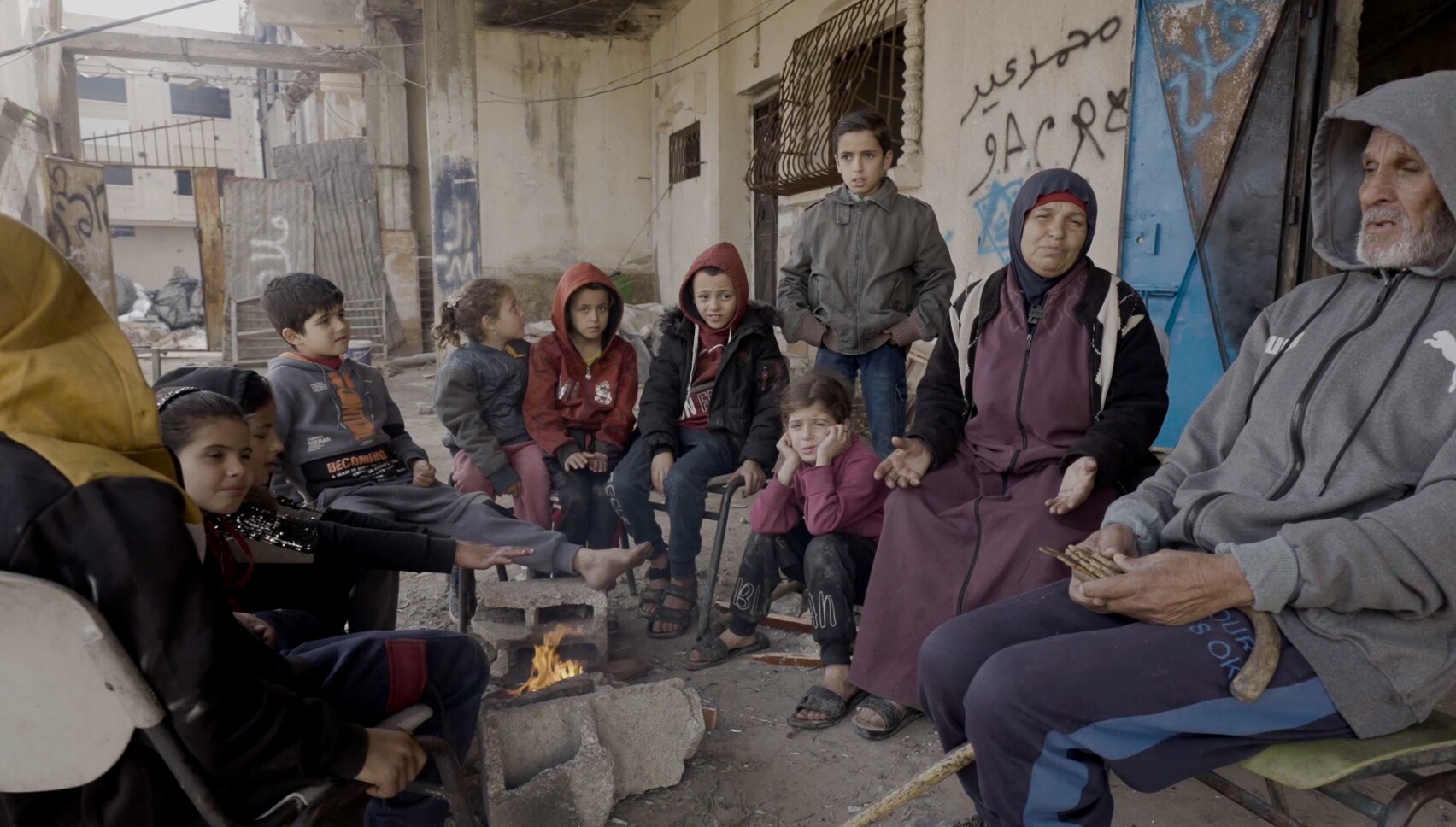 A woman and a man children sit gathered around a fire with their children in the rubble of a damaged home.