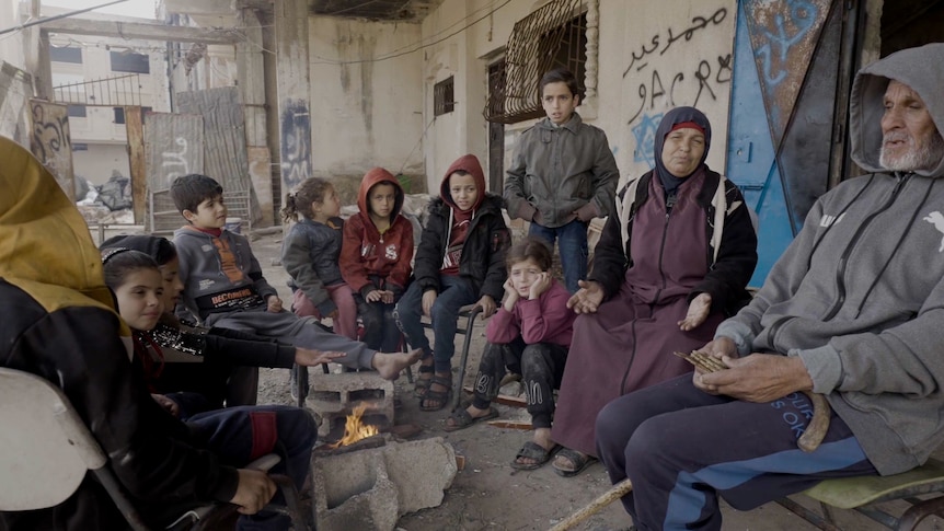 한 여성, 한 남성, 아이들이 폐허가 된 집의 잔해 아래에서 아이들과 함께 불 주위에 옹기종기 모여 앉아 있습니다.