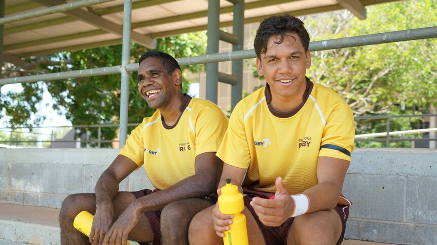 Two Indigenous teenage boys sit in the grandstand and have a drink.