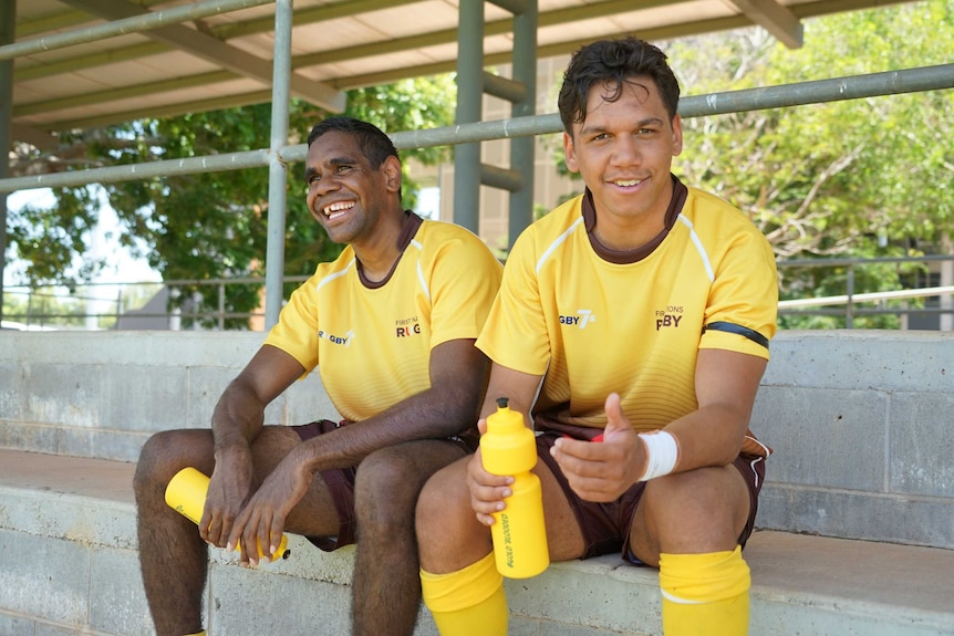 Two Indigenous teenage boys sit in the grandstand and have a drink.