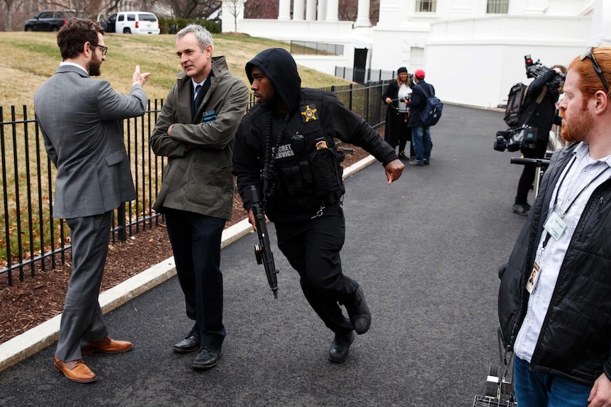 A Secret Service officer holds a gun as he runs past two men in suits chatting by a fence