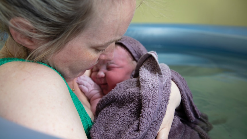 close of up blonde woman with baby wrapped in purple after home birth with birthing pool in background