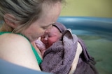 close of up blonde woman with baby wrapped in purple after home birth with birthing pool in background