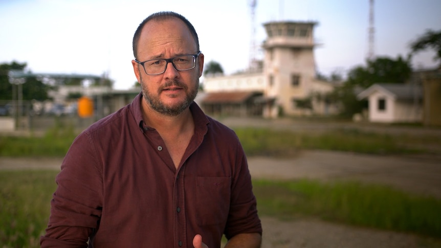 Mark Willacy delivers a piece to camera an air control tower in the background.