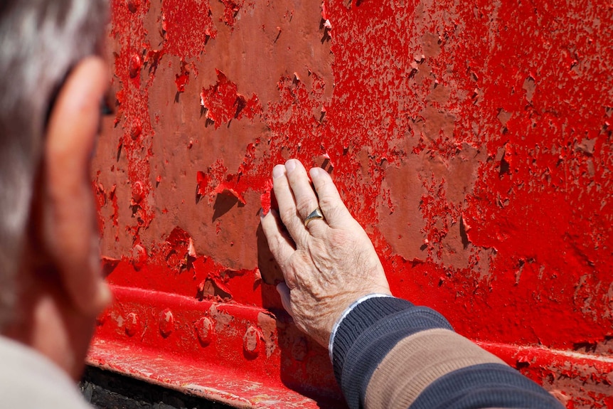 A man's hand touching peeling red paint on a boat's red hull.