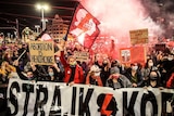Hundreds of people are seen protesting in a Wroclaw street at night with red smoke above them. Several hold placards.
