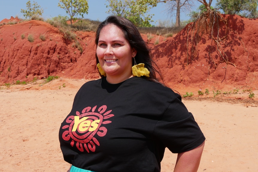 A woman standing on the beach and smiling while wearing a t-shirt with the word 'yes' printed in black, yellow and red.