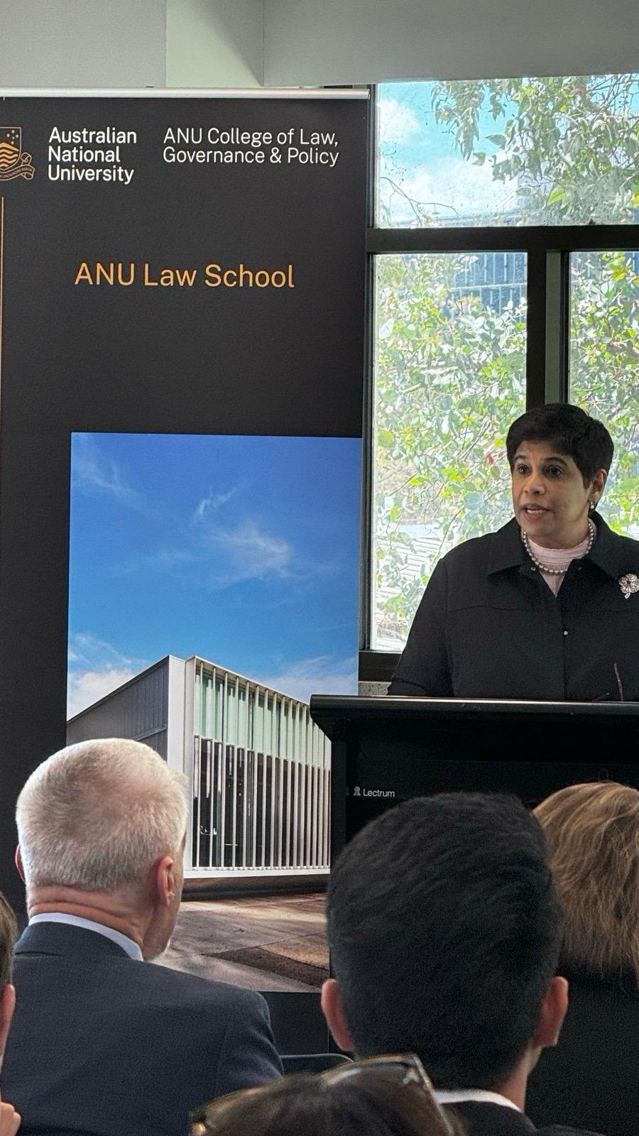 A woman with short black hair stands behind a lectern speaking to people out of frame