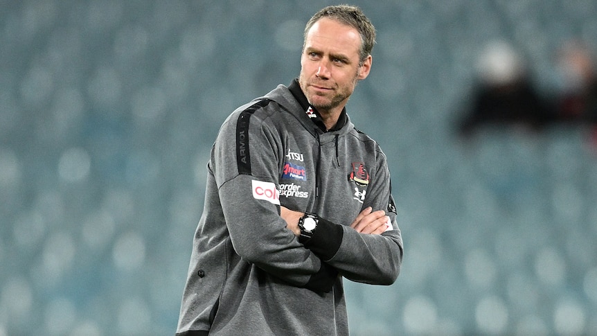 Essendon AFL coach Ben Rutten stands on the MCG with his arms crossed.