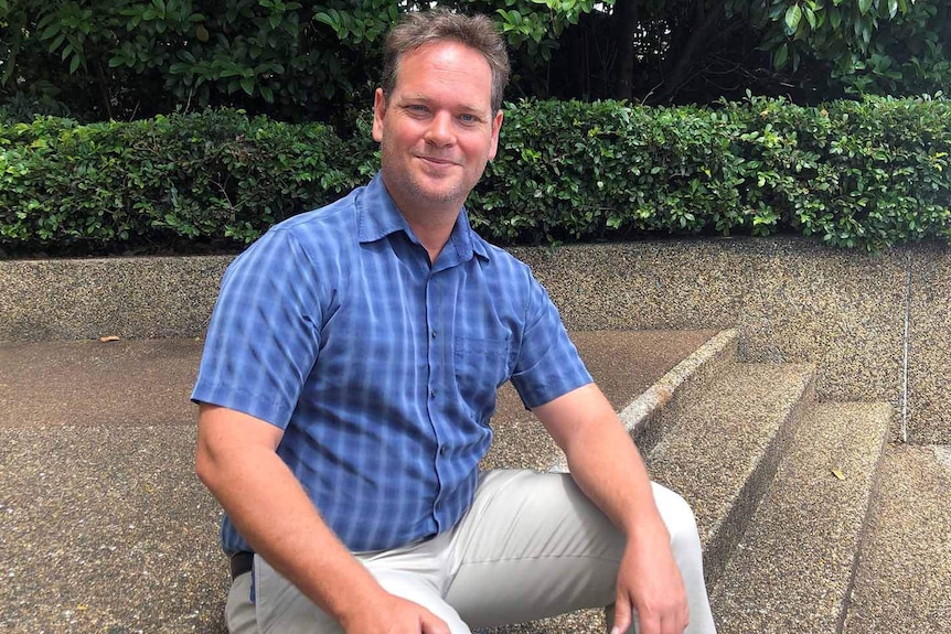 A smiling man dressed in smart casual clothes sitting on steps