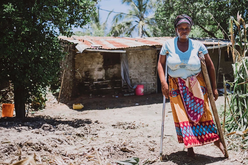 A woman leaning on mismatched, rudimentary crutches, standing in front of a shack on a sunny day.