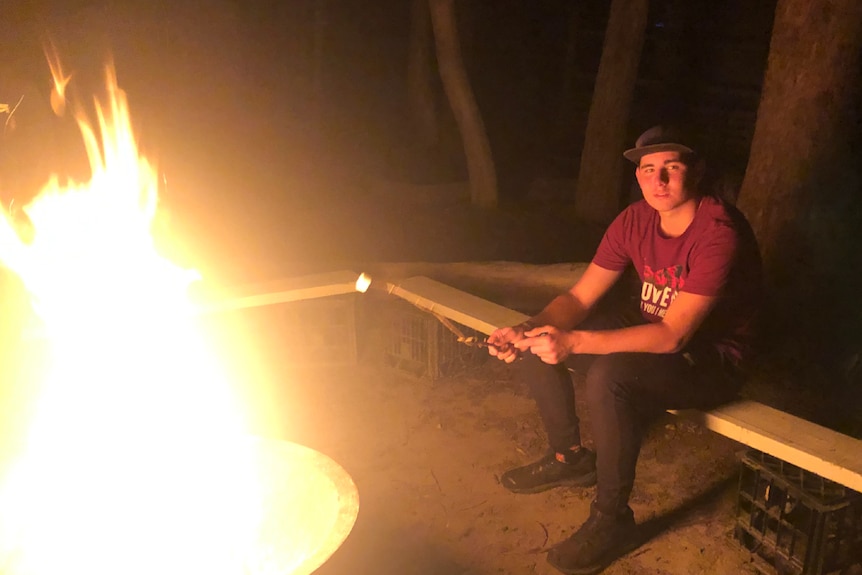 Teenage boy sits beside a fire.