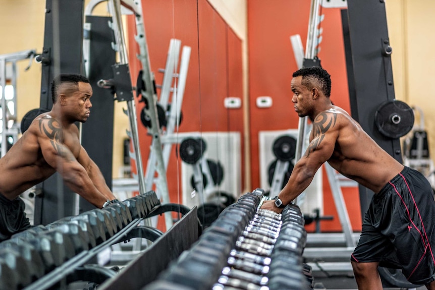 A shirtless male athlete leaning over a weights rack at the gym looking at himself in the mirror