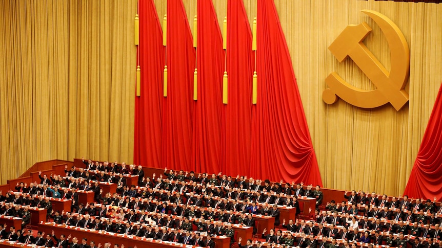 Rows of men sitting down raise their hands in a great hall with the communist symbol behind them.