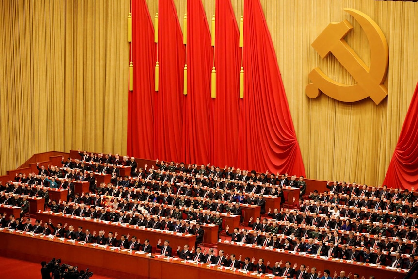 Rows of men sitting down raise their hands in a great hall with the communist symbol behind them.