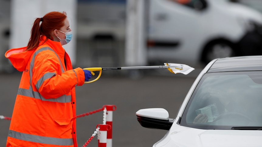 A health worker in orange attire collects a COVID-19 test from a car