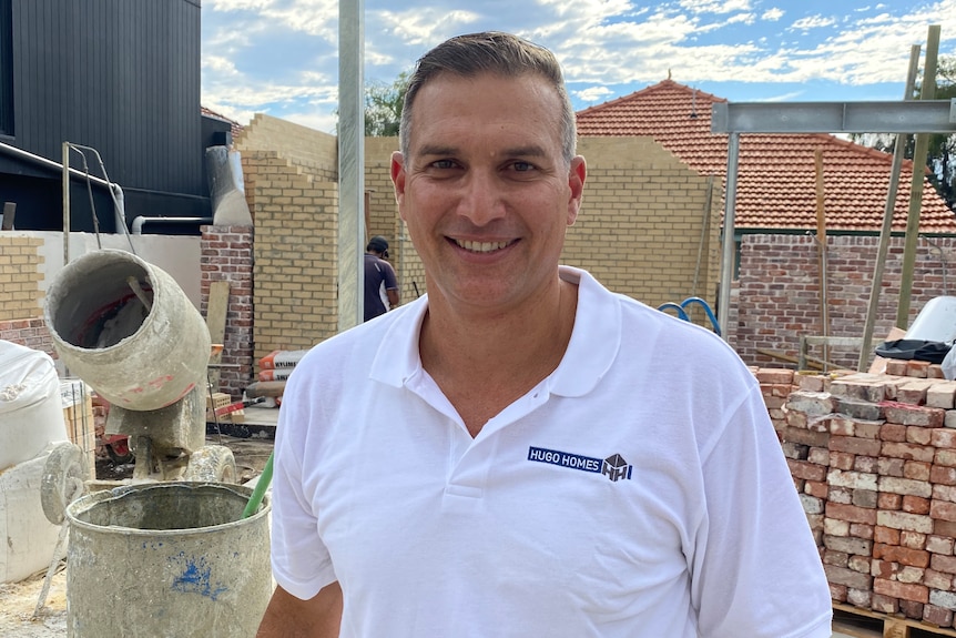 A man wearing a white polo shirt stands in a building site. There are bricks and a cement mixer behind him