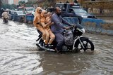 A South Asian man in blue rides a motor bike on a flooded street with three girls in matching Islamic garb sitting behind him