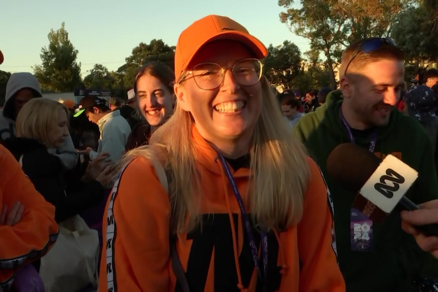 A young woman smiling while wearing an orange jumper and hat with the McLaren logo on it.