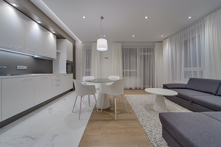 The interior of a contemporary apartment with white/grey kitchen, table, curtains and ceiling, pale timber floor and dark grey 