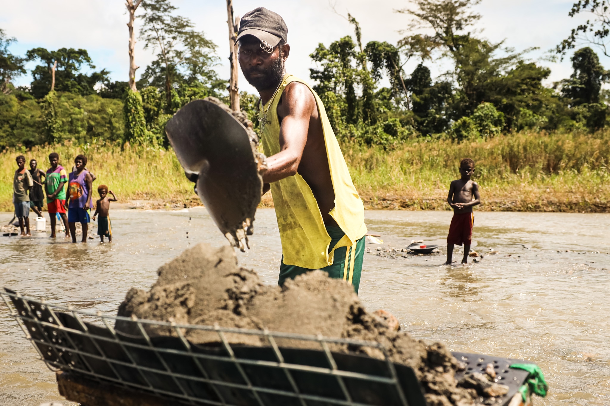 A Black man shovels dirt into a pile while kids watch on from nearby.