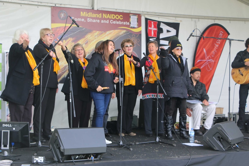 About a dozen members of the Madjitil Moorna Choir singing on stage wearing yellow scarves with a NAIDOC banner behind them.