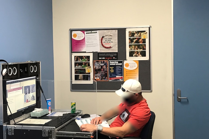 A man in a red t-shirt sits at a computer in a small office. His face and tattoos on his upper arms are blurred.