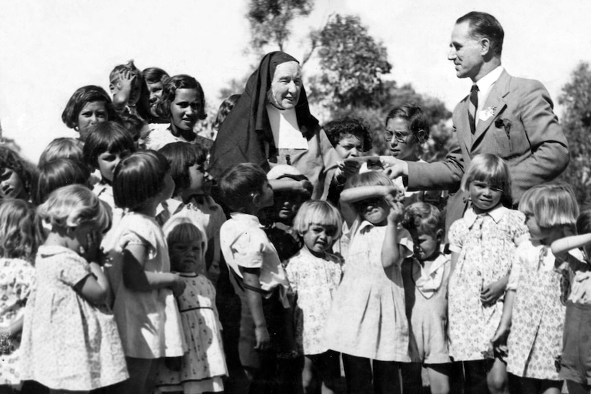 A black-and-white photo shows a nun surrounded by children, receiving a cheque from a man in a suit.