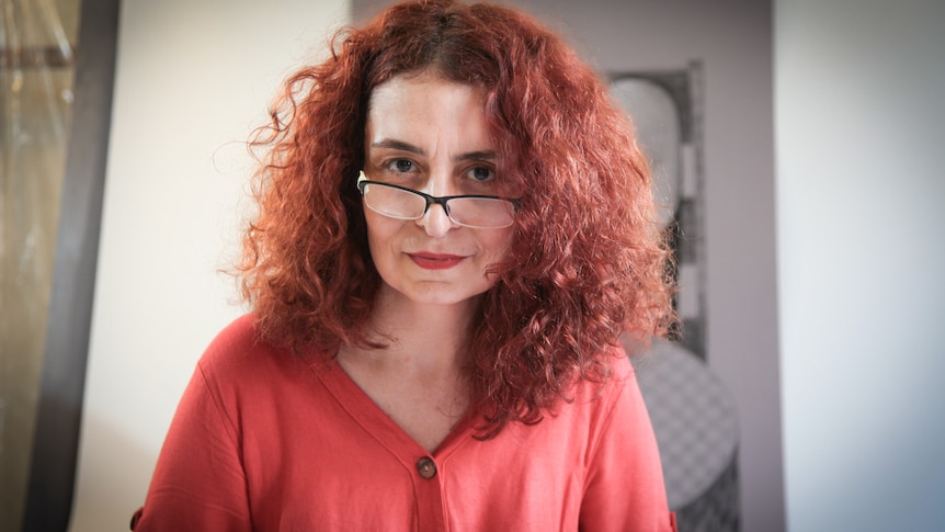 A woman with red hair and wearing glasses and a red dress poses for a photo.