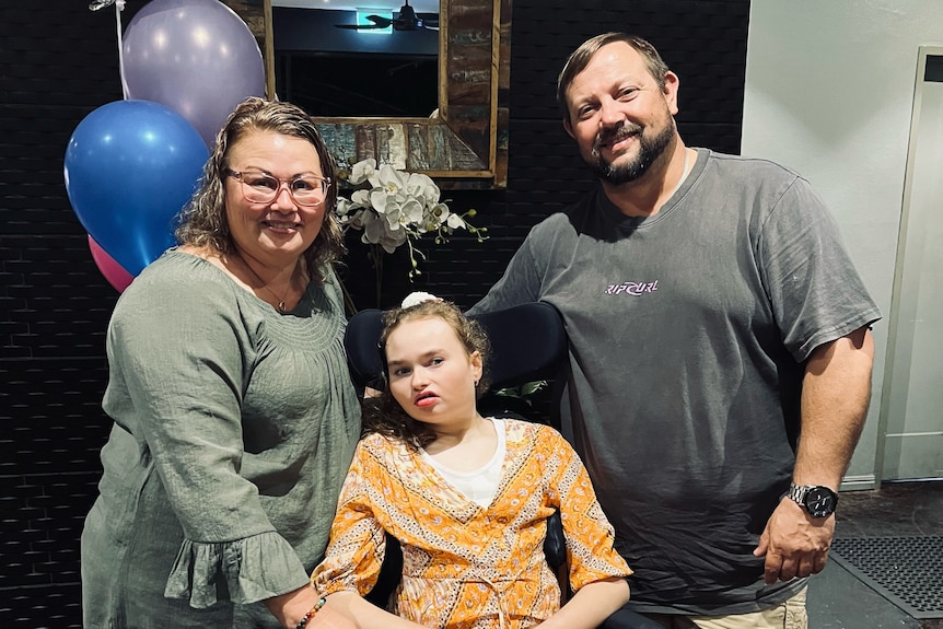 A teenage girl in a wheelchair with her parents with baloons in the background.