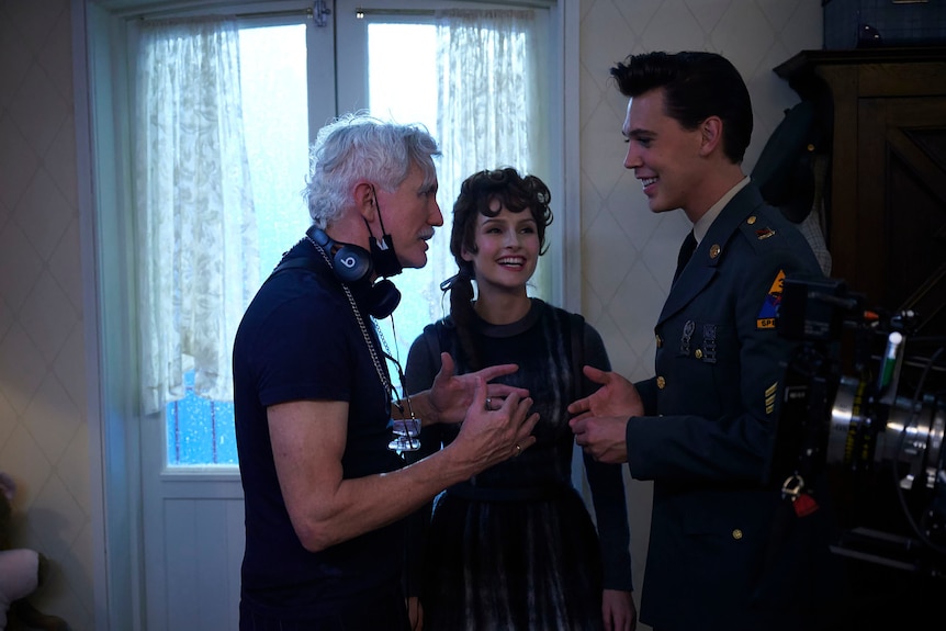 A male director with white hair is giving notes to an actress with 50s style hair and dress and an actor dressed as Elvis on set