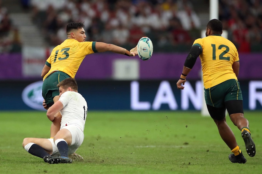 A Wallabies player gets a one-handed pass away to his teammate on the right as he is tackled by an England opponent.
