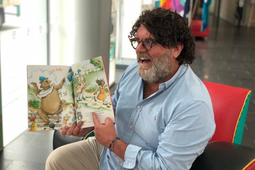 Russell Woolf sitting inside holding an open children's book and laughing.