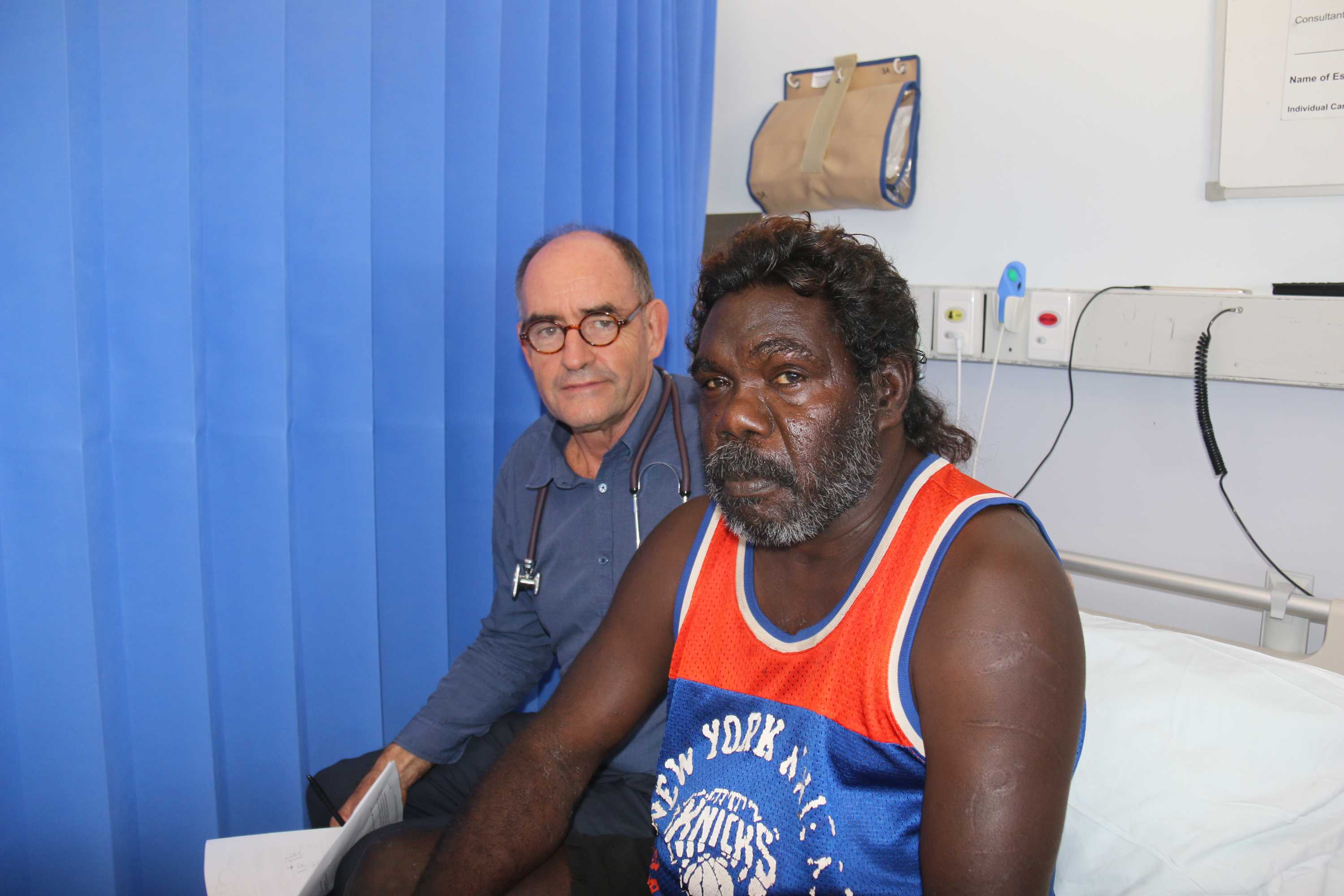 Two men sit together on a hospital bed.