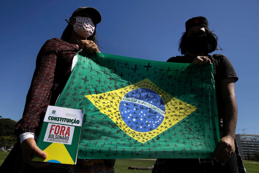Two protesters wearing face masks hold up a Brazilian flag marked with black crosses