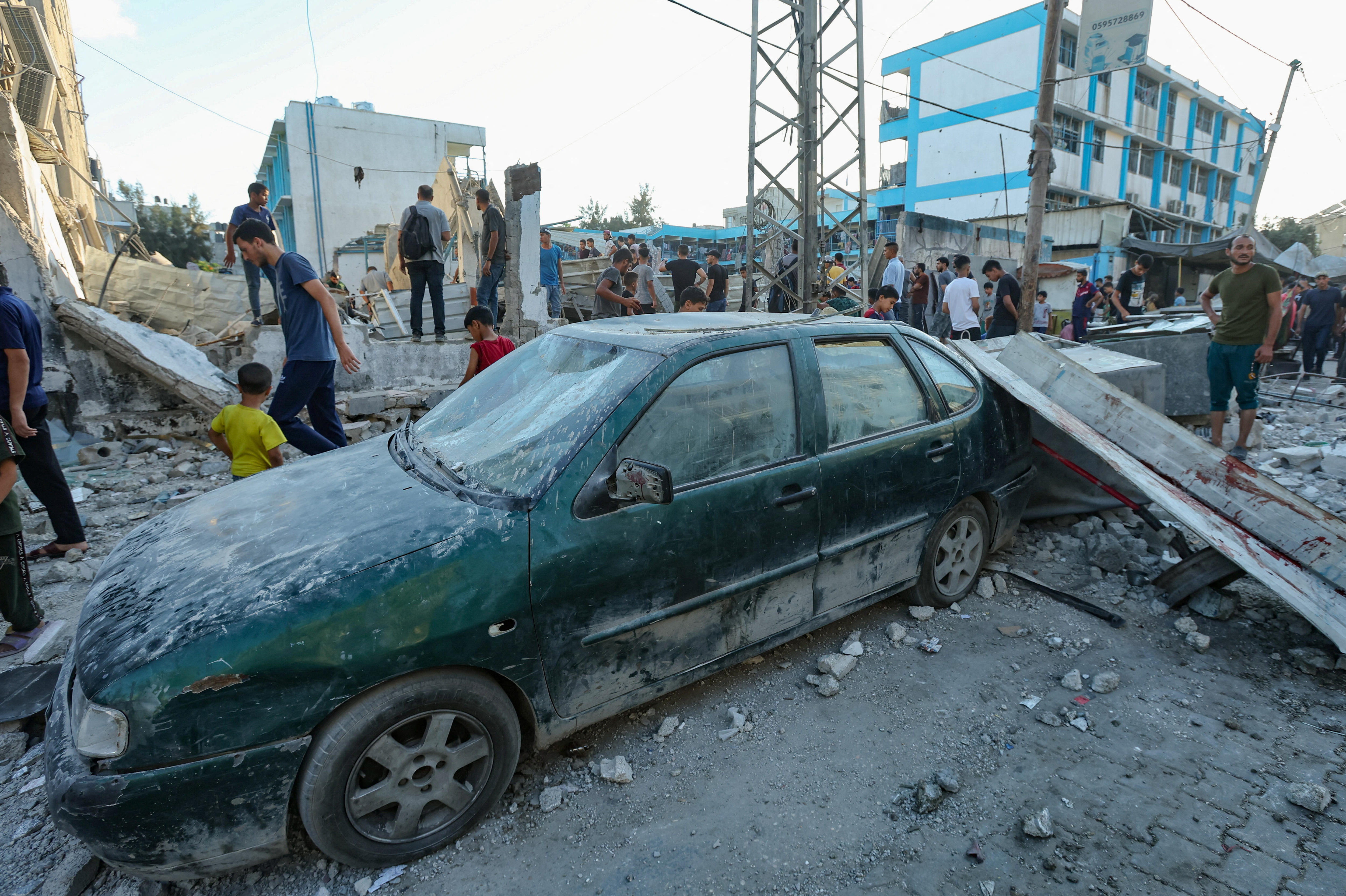Rubble lies across a damaged green car as crowds inspect the scene of an air strike