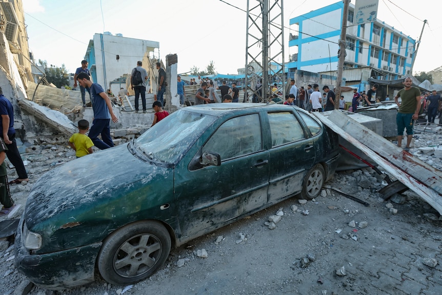 Rubble lies across a damaged green car as crowds inspect the scene of an air strike