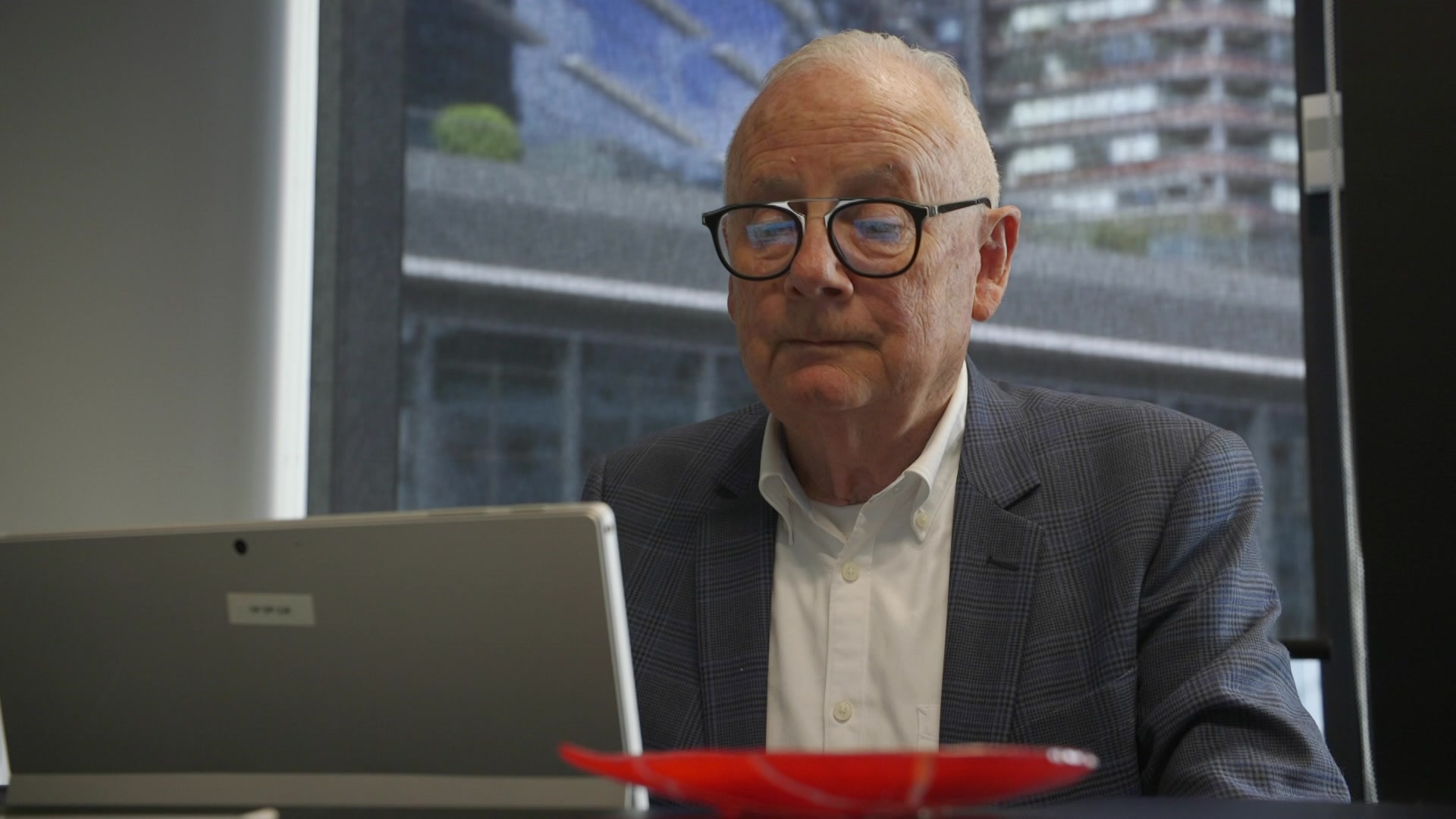 A man with white hair and glasses sits in front of a computer looking down at the screen