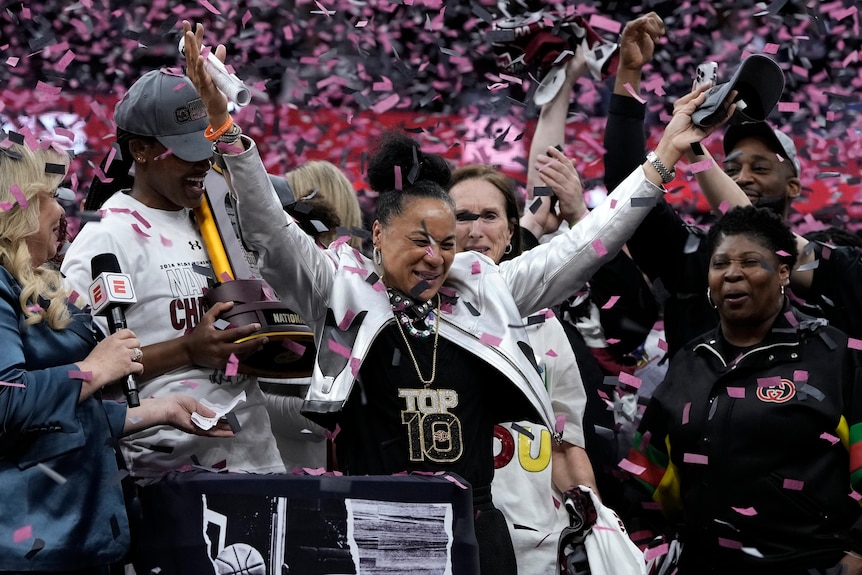 A women's basketball team is showered in confetti standing on a podium, after winning a championship  