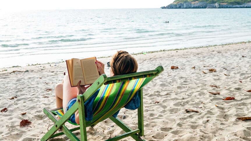 A woman reads a book in a deck chair on a beach.