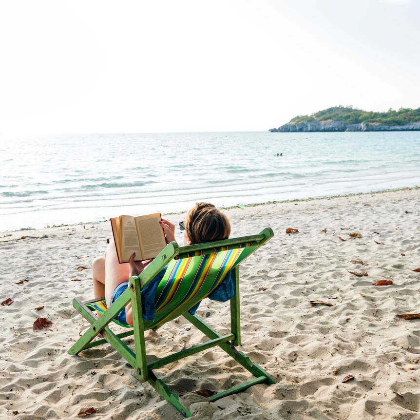 A woman reads a book in a deck chair on a beach.