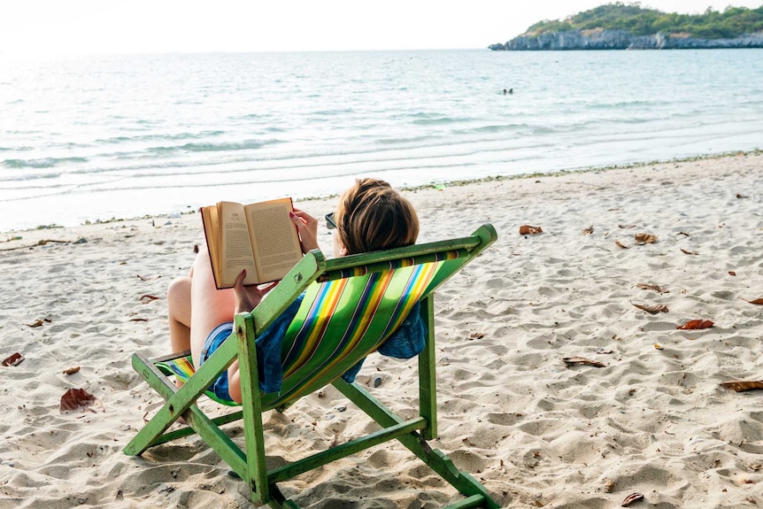 A woman reads a book in a deck chair on a beach.