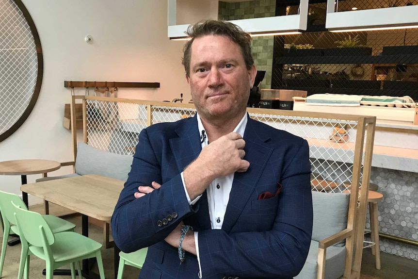 A portrait of a man, Chris Shine, with his hand at his shirt collar inside an empty cafe.