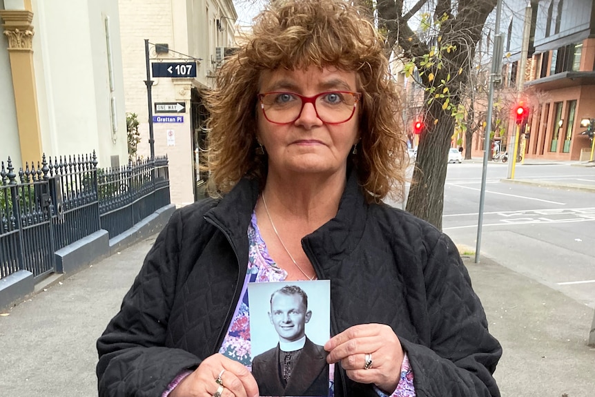 A woman with brown curly hair and red spectacles poses on a city street holding a black and white photo of a priest