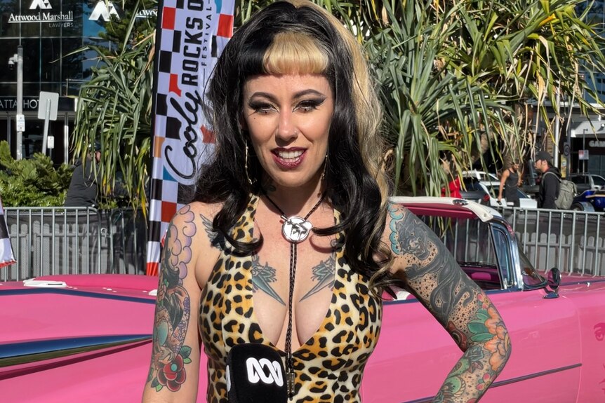 A woman dressed in leopard print with blonde and black hair standing in front of pink Cadillac